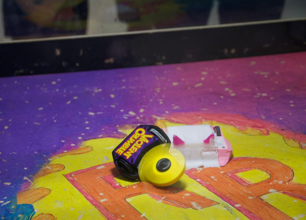 VIOLENT CRUMBLE: IN ACTION! A close up in an area, where small ant-weight class robots are battling, primarily 3D Printed. Violent Crumble (purple robot with a candy wrapping looking title, with a thick yellow plastic blade spinning like crazy), taking on another robot that is a ramp