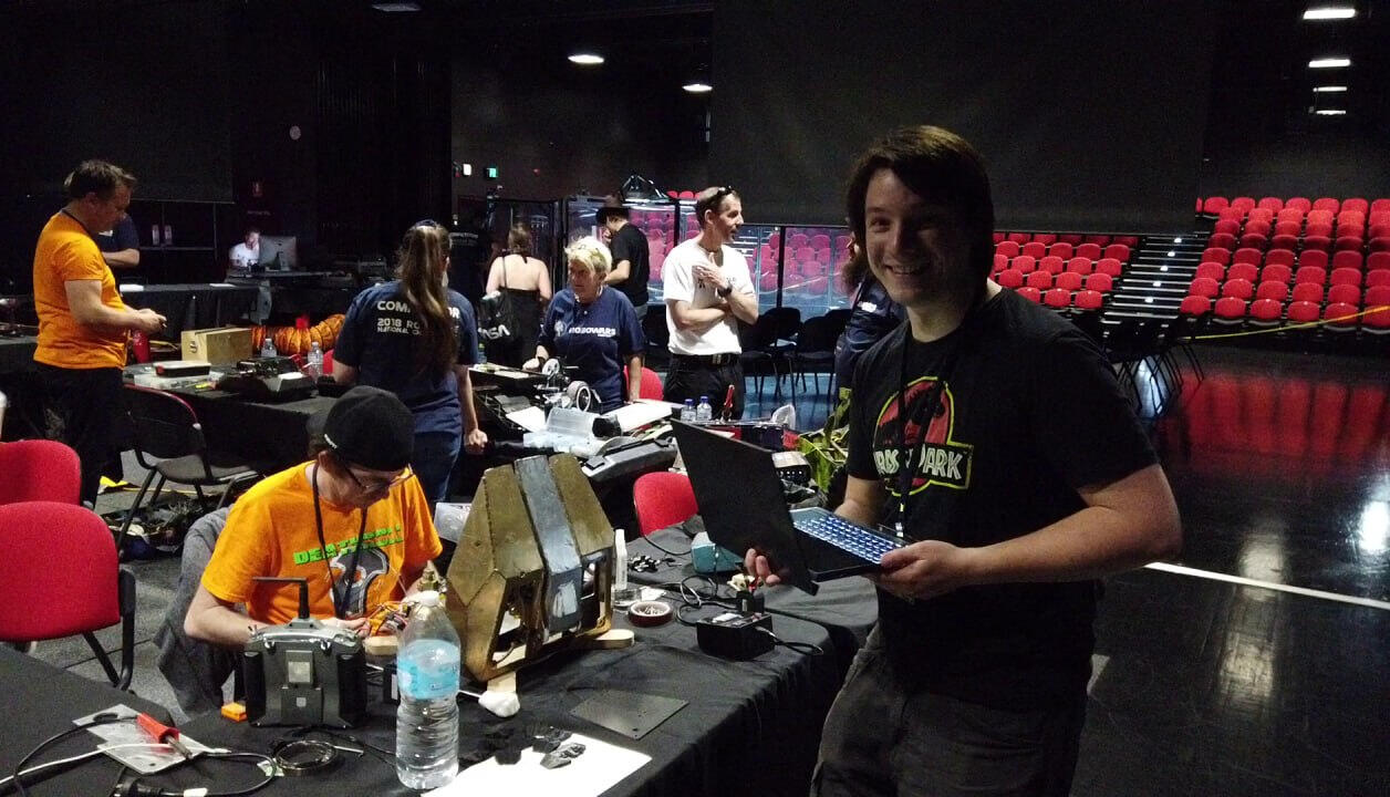 Robot Wars: 2018! Angus Deveson at a past Robot Wars Competition in Australia: Standing to the side next to the competitors work stations while they set up and tend to their robots, smiling and dressed in a Jurassic Park black shirt, holding a laptop.