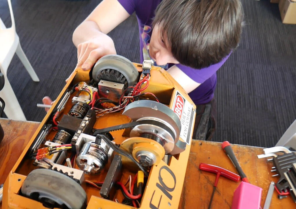 Quick patch up job! Angus Deveson at a robot event: close up shot of Angus peering into a large metal combat robot (Beetle-weight class), examining the wheels. Predominantly metal with a tidal wave-looking thick spinner blade weapon.