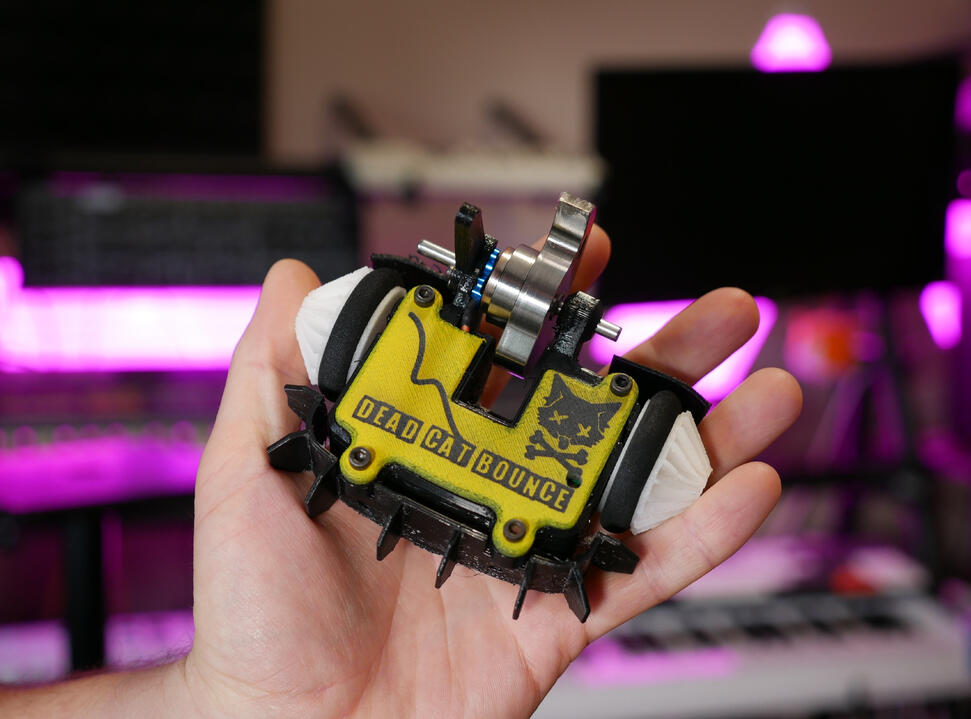 DEAD CAT BOUNCE: 2025 Angus's hand holding up small combat robot in his studio: Ant-weight class, with a metal spinner blade custom made. 3D printed yellow cover with a black logo "Dead Cat Bounce" on it. Has a graphic design of a goofy black cat with "x" eyes and cross bones.