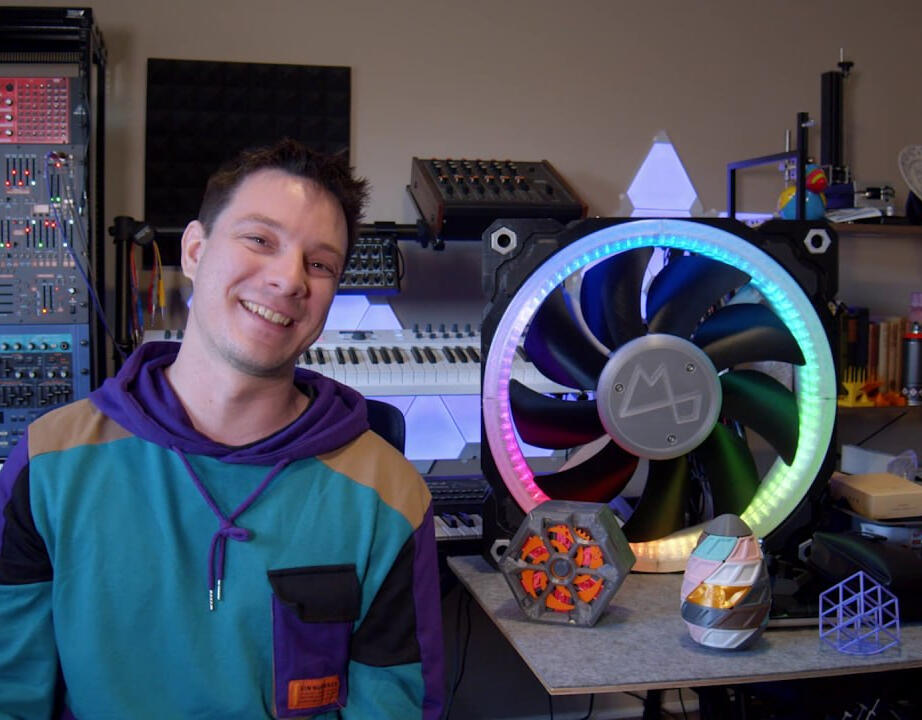 Angus Deveson and his various projects! Angus Deveson in his workshop, standing beside his 3D printed projects which is on a table to the right. Featuring: a giant 3D printed RGB fan, a 3D printed Makers muse puzzle box, and a 3D printed puzzle box that looks like an egg.