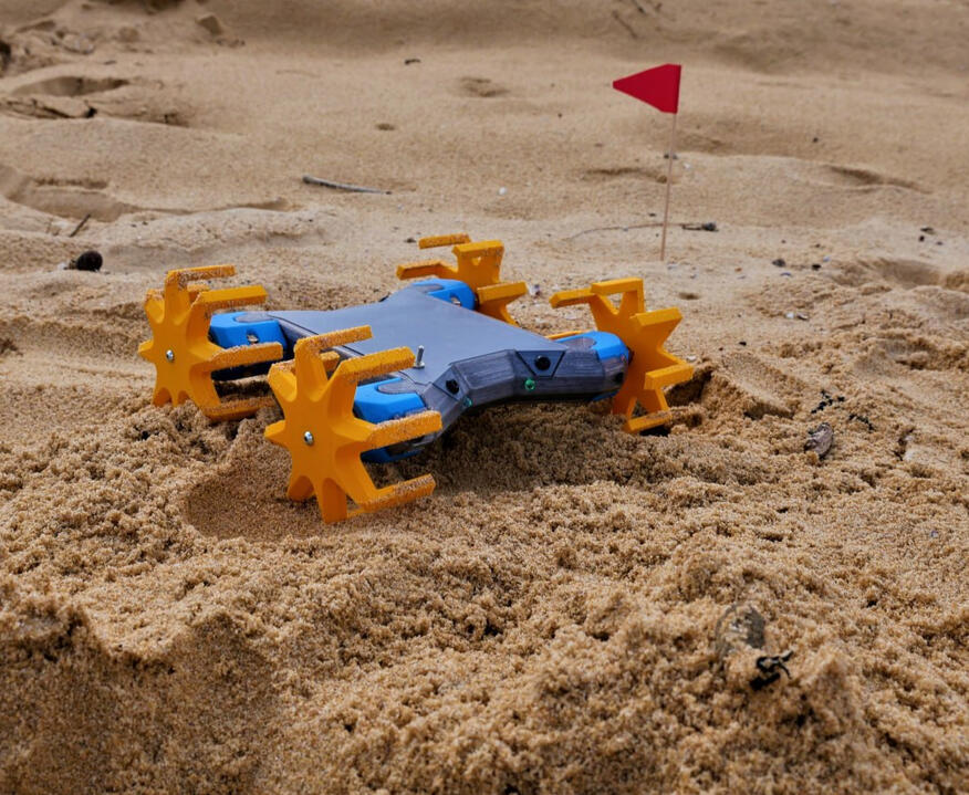 Sand-Track Off Roader Makers Muse's Off-Roader Vehicle, primarily 3D Printed! It's a small blue robot/vehicle with 4 spindly looking orange wheels, the vehicle navigating over the beach sand around a tiny red flag sticking out of the sand.