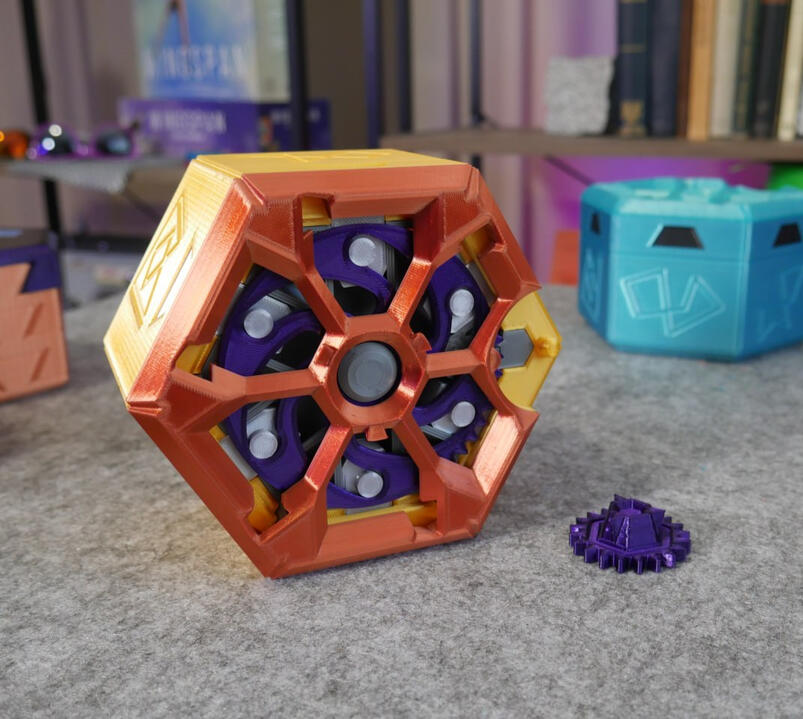 Expanding Puzzle Box! A close up look at Makers Muse's 3D printed expanding puzzle-box design on a gray surface: Hexagon shaped and coloured orange and purple. Many mechanisms and gears are evident in the design.